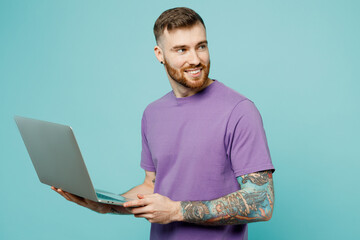 Young smiling happy IT man he wears purple t-shirt hold use work on laptop pc computer look aside...