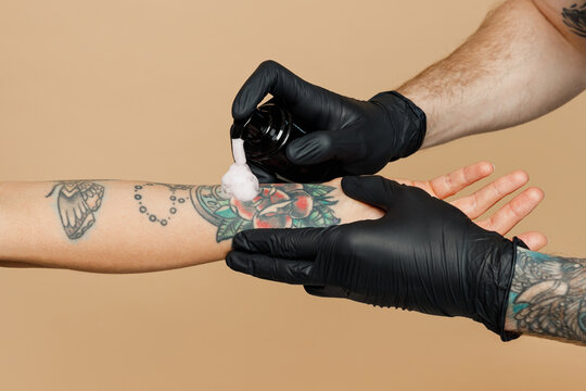 Cropped close up photo shot of tattooer master artist tattooed man wearing black sterile gloves apply cream on female hand after making colored tattoo art on body isolated on plain beige background.