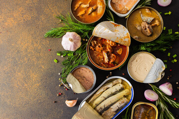 Different open tin cans with canned fish among spices and herbs on a brown background, canned salmon and mackerel, sprat and sardine, tuna and herring and fish pate, top view