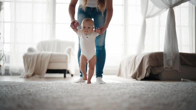 Mom holding hands baby who trying to walk in living room at home. Little cute baby doing first steps with mother's help. Childhood, babyhood, motherhood, babycare, parenting, maternity leave concept.