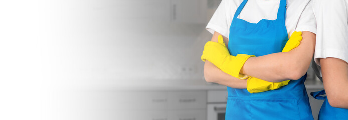 Cleaning team of two people with their arms crossed on their chests against the background of a blurry kitchen. Professional female workers in aprons and yellow rubber gloves cleaning company. Banner