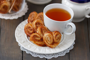 Homemade French Palmier cookies on white plate with a cup of tea and teapot on background. Ready to eat sugar hearts pastries made from puff pastry.  Selective focus, horizontal