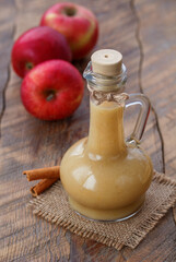 Smooth homemade applesauce in a glass jug with fresh red apples and cinnamon stiks on a wooden table. Selective focus, vertical.