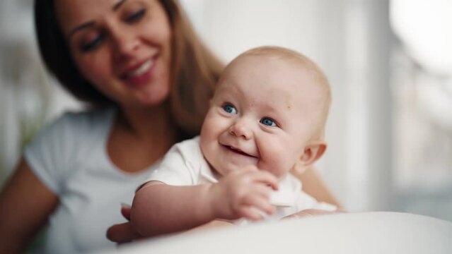 Happy mom babysitting little baby smiling talking at home in maternity leave. Motherhood babycare concept. Mother looking at her positive laughing kid. Babyhood, childhood, parenting, maternity leave.