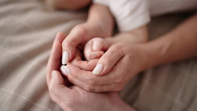 Close-up view of sleeping in bed baby's little hand in mom and dad's hands. Family love and babycare childcare, parenthood childhood concept. Mother, father and newborn child lying resting together.