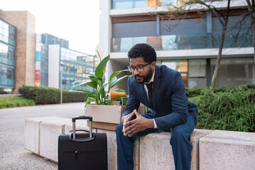 Depressed businessman with suitcase and his belongings sitting outside office building