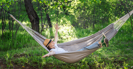 Young happy caucasian woman in a hat lying in a hammock in a green garden enjoying a summer day - 593531177