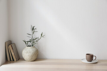 Olive tree branches in beige textured vase. Brown cup of coffee, tea and old books on wooden table. White wall background. Minimalistic Scandinavian interior, dinning room. Summer breakfast.