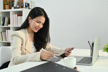Attractive asian woman small business entrepreneur holding smart phone and using laptop at modern workplace
