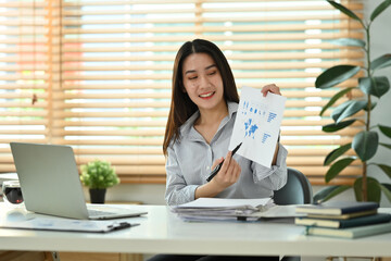 Smiling young businesswoman holding financial graph reports making virtual online presentation via laptop computer