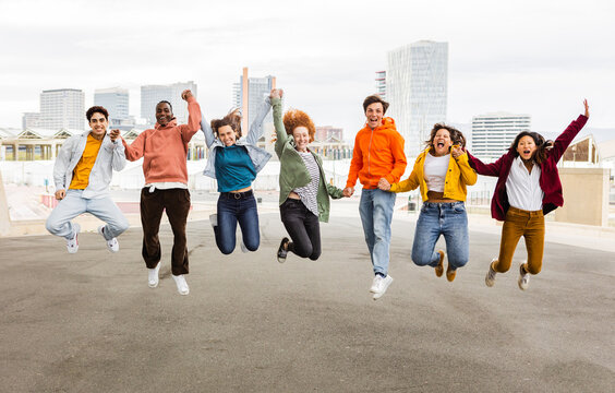 Diverse happy group of young friends celebrating having fun outdoors. Community concept with teenage people jumping holding hands over city urban background