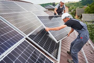 Men workers mounting photovoltaic solar moduls on roof of house. Electricians in helmets installing solar panel system outdoors. Concept of alternative and renewable energy.