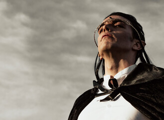 Brown-haired man with elf ears, tiara on head and black cape, looks forward