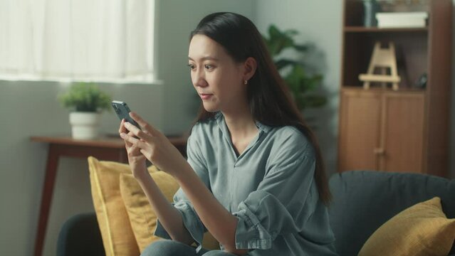 Beautiful young Asian woman hand holding mobile phone playing social media. Happy attractive girl smile looking at smartphone browsing internet chatting with friends sit on sofa in cozy living room