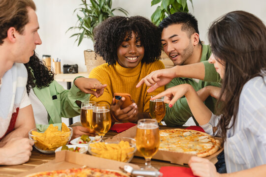 laughing friends eating pizza and having fun. They are enjoying eating and drinking together. they look at the smart phone laughing watching videos of social networks
