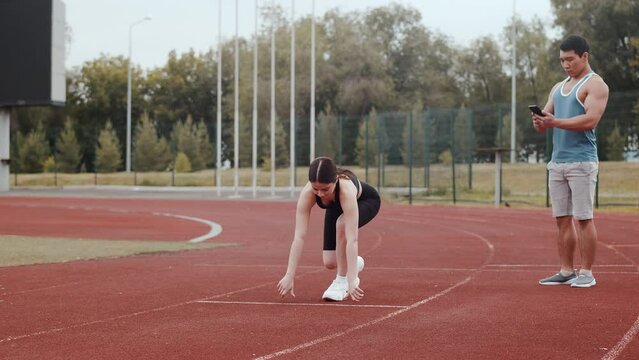 Young caucasian female athlete starts running from a low-start position while his asian trainer controls the time at the running track in an outdoor stadium