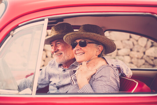 Youthful senior old couple enjoy travel with classic red car together having fun and love. Man hugging woman inside automobile vehicle during summer tourism. Happiness and retirement lifestyle people.