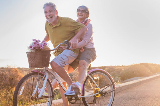 Youthful and playful happy senior old couple enjoy outdoor leisure activity riding together the same bike. Man carrying aged woman in healthy active lifestyle. Retiremd people using bicycle