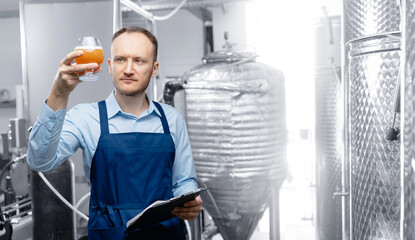 Inspector checking drink in alcohol brewery manufacturing factory. Worker examining quality of craft beer at brewery in wineglass