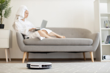 White automatic robot vacuum keeping carpet dust-free while female owner relaxing with portable computer on cozy couch indoors. Wireless tech gadget providing healthy environment in airy apartment.
