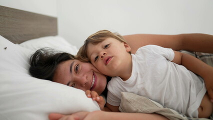 Obraz na płótnie Canvas Mother and son laid in bed in embrace looking at camera smiling. Motherhood lifestyle child and mom together