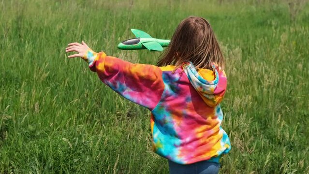 Preteen girl child wearing colorful hoodie playing with toy plane in the field with green grass in sunny day and smiling. Pretty young female kid launching airplane at the nature and has fun