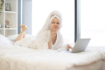 Beautiful young lady in white after-shower robe daydreaming on cozy bed while running portable computer in sunny room of modern apartment. Charming female adult enjoying comfortable lifestyle.