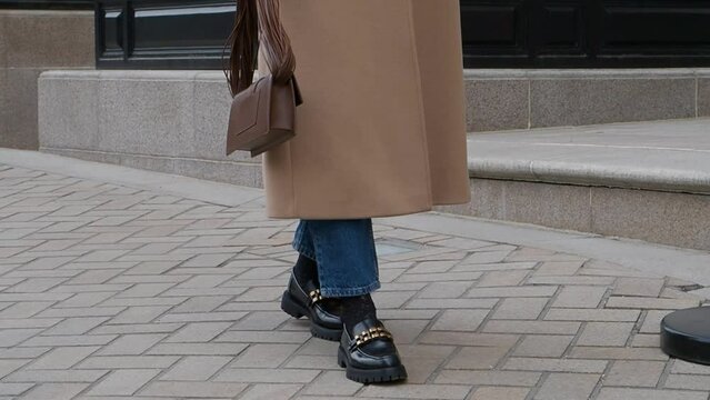 Woman walking street in fashionable spring or autumn clothes cashmere coat, jeans, black loafers shoes and handbag. Female model in motion, street style fashion, close up legs
