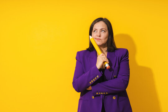 Middle aged woman wearing purple blazer is holding giant pencil on yellow background