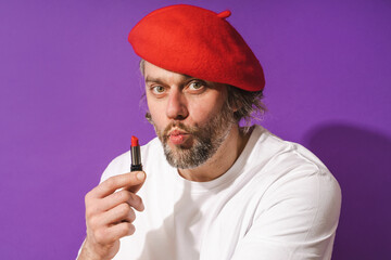 Funny middle aged man wearing red beret is holding lipstick in his hand against purple background