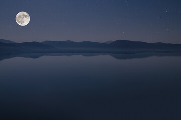 Silhouette of mountains near lake on the huge full moon and night stars sky background.