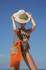 Young woman in tropical printed swimwear and straw hat cary orange crochet tote bag empty sandy beach on, summer fashion style, relaxed vacation	