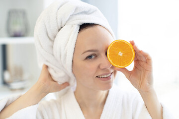 Close-up view of young healthy female wearing soft towel and holding orange fruit piece over her eye on blurred background of bright room. Happy lady achieving natural beauty effect of skin glow.