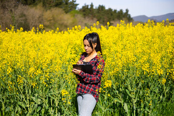 Naklejka na ściany i meble Agricultural woman technician with a folder checking the growth of rapeseed crop yellow flower. Female agricultural engineer working in field. Brassica napus crop. Modern technologies in agriculture.