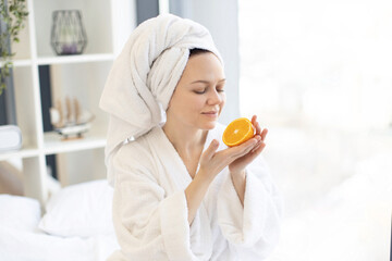 Serene healthy female after bath taking pleasure of fresh citrus fruit aroma while relaxing in peaceful bedroom interior. Charming adult person using rich in vitamin C orange for high-end beauty life.