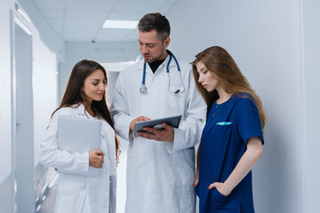 The male chief physician shows the patient's diagnosis to his colleagues at the clinic on a tablet. Three colleagues are talking in the hallway.