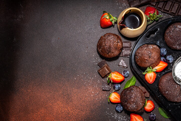 Chocolate muffins with berry. Baking muffin pan with homemade sweet dark chocolate cupcakes with cocoa sauce, sugar powder and fresh strawberry, blueberry, on dark brown background copy space