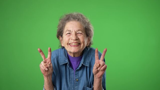 Portrait of happy elderly senior old woman with wrinkled skin and grey hair smiling and giving peace or victory sign gesture with hands isolated on green screen background.