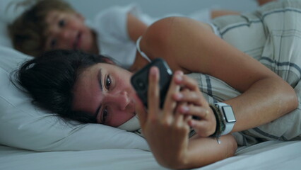 Fototapeta premium Woman lying in bed looking at phone in the morning. Candid lifestyle mother checking cellphone screen. People addicted to technology