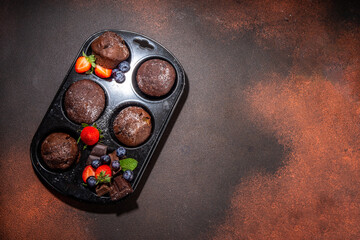 Chocolate muffins with berry. Baking muffin pan with homemade sweet dark chocolate cupcakes with cocoa sauce, sugar powder and fresh strawberry, blueberry, on dark brown background copy space