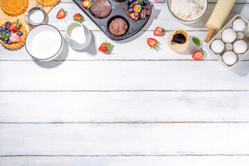 Summer baking background with baked pastry - berry cakes, muffins, mini pie tarts, with cooking baking ingredients, flour, eggs, rolling pin, cream milk, sugar, strawberry, white wooden background