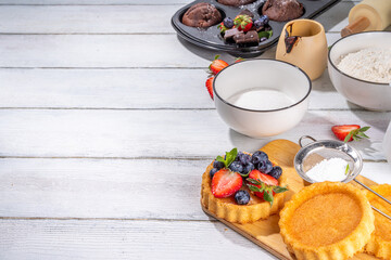 Summer baking background with baked pastry - berry cakes, muffins, mini pie tarts, with cooking baking ingredients, flour, eggs, rolling pin, cream milk, sugar, strawberry, white wooden background