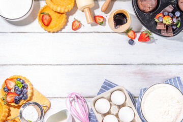 Summer baking background with baked pastry - berry cakes, muffins, mini pie tarts, with cooking baking ingredients, flour, eggs, rolling pin, cream milk, sugar, strawberry, white wooden background