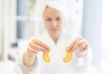 Focus on pair of golden eye patches being held by young caucasian woman wearing bathrobe and soft towel on head in home interior. Anti-aging under-eye masks fighting dark circles and fine lines.