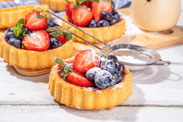 Sponge mini tart cakes with summer berry. Portioned vanilla dessert cups with fresh strawberry, blueberry, raspberry, on white wooden background copy space