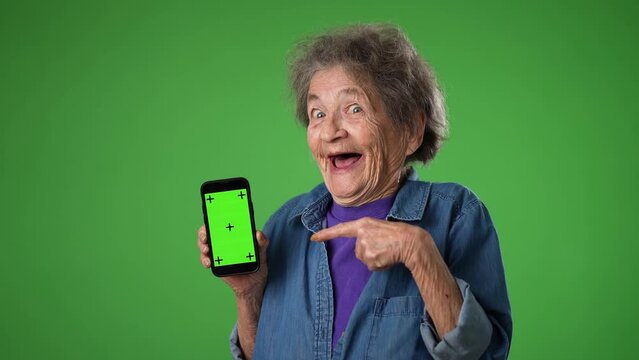 Funny crazy elderly old toothless woman points to phone with green screen tracking points isolated on green screen background with mock up copy space. Hold mobile phone with blank empty screen