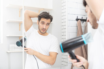 Man sweating excessively and trying to dry his armpits with hair dryer in front of mirror in the...