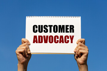 Customer advocacy text on notebook paper held by 2 hands with isolated blue sky background. This message can be used as business concept about customer advocacy.