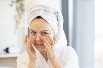 Close up view of charming adult person in white bathrobe posing with paper sheet mask on face while relishing skincare beauty therapy at home. Caucasian lady making use of facial cosmetics in bedroom.