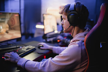 Side view of young gamer in headphones playing computer game in cyber club. - obrazy, fototapety, plakaty